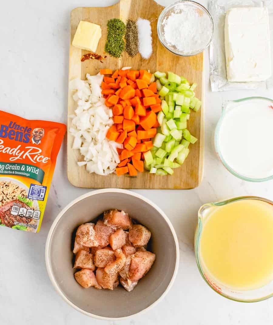 Ingredients for the chicken and wild rice soup including diced onion, carrots, and celery on a small wooden cutting board, raw chicken, milk, cream cheese, chicken broth, and rice.