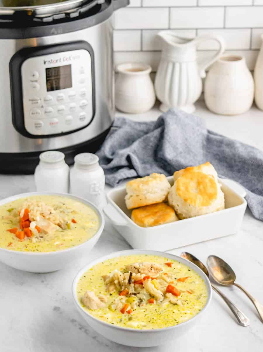 Two bowls of chicken and wild rice soup in white bowls next with two spoons, some biscuits in a white rectangle serving dish, and an Instant Pot in the background.