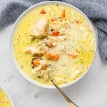 A bowl of creamy instant pot chicken and wild rice soup filled with chunks of chicken, carrots, and other vegetables. A spoon rests in the bowl, and a blue patterned cloth is partially visible next to it, all set on a light gray surface.