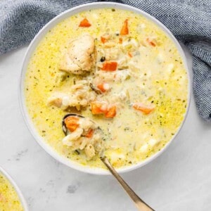 A bowl of creamy instant pot chicken and wild rice soup filled with chunks of chicken, carrots, and other vegetables. A spoon rests in the bowl, and a blue patterned cloth is partially visible next to it, all set on a light gray surface.