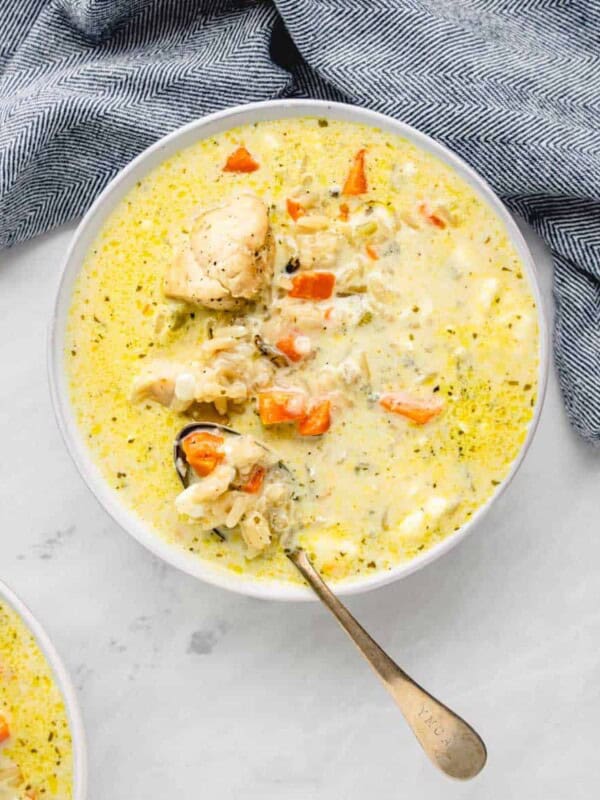 A bowl of creamy instant pot chicken and wild rice soup filled with chunks of chicken, carrots, and other vegetables. A spoon rests in the bowl, and a blue patterned cloth is partially visible next to it, all set on a light gray surface.