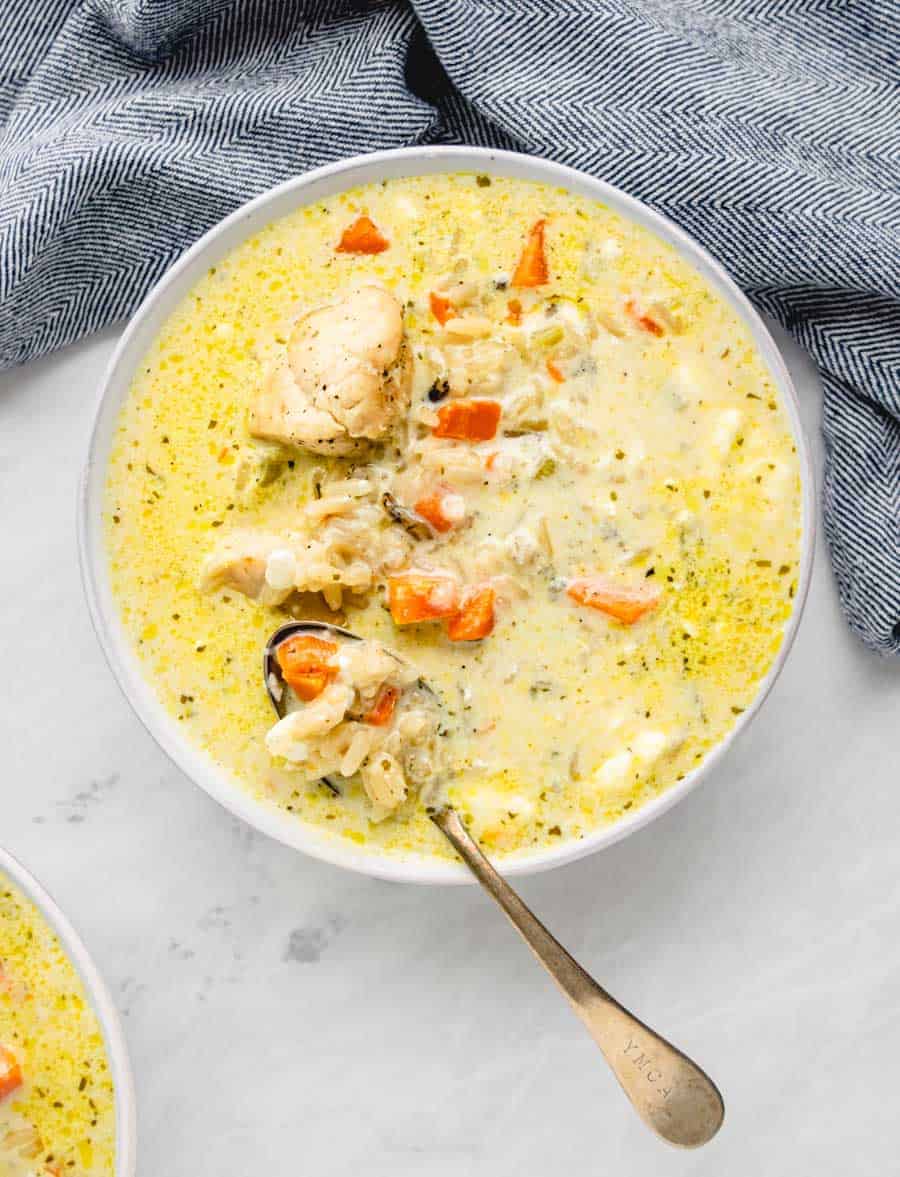 A bowl of creamy instant pot chicken and wild rice soup filled with chunks of chicken, carrots, and other vegetables. A spoon rests in the bowl, and a blue patterned cloth is partially visible next to it, all set on a light gray surface.