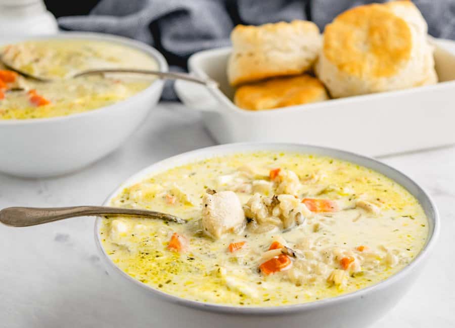 A bowl of chicken and wild rice soup in a white bowl with another bowl of the soup and some biscuits in a white rectangle serving dish in the background.