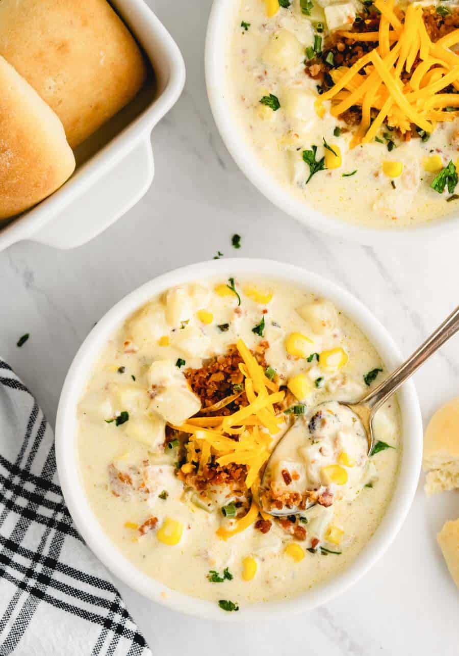 A bowl of potato topped with bacon crumbles and cheese with rolls in the background.