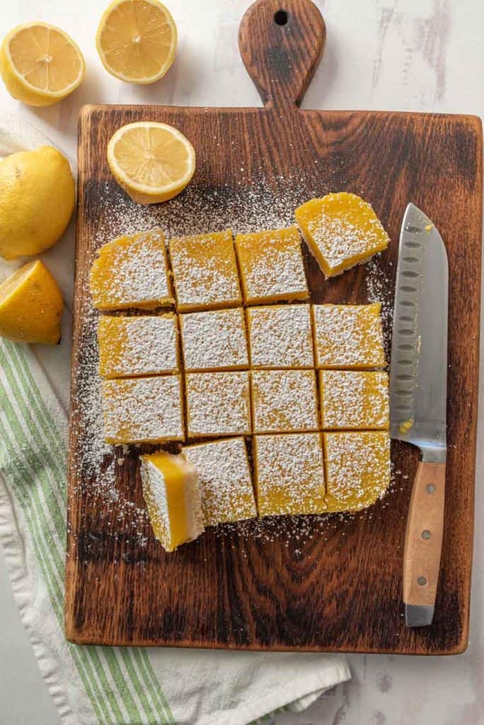 top view of lemon bars on wood cutting board with knife and lemons around it