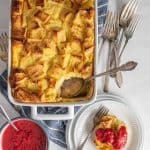 top view of French toast breakfast casserole dish with a portion of it on a plate and a bowl of raspberry sauce