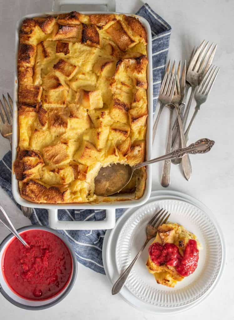top view of French toast breakfast casserole dish with a portion of it on a plate and a bowl of raspberry sauce