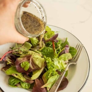 A hand pours poppy seed dressing from a jar onto a plate of mixed green salad with a fork resting on the plate, offering inspiration for how to make salad dressing at home.