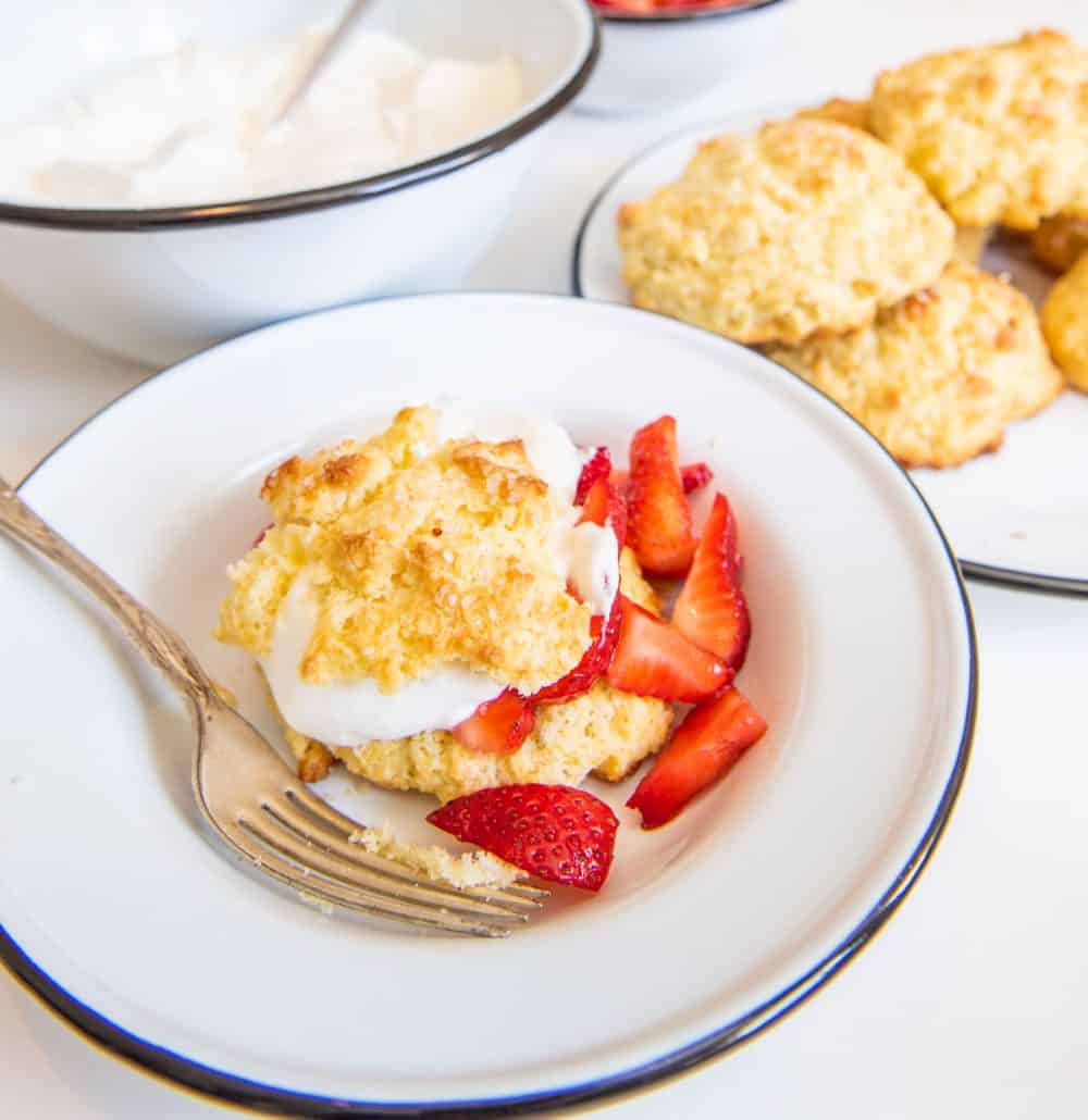 A bowl with a strawberry short cake biscuit, whipped cream, and sliced strawberries with a fork and short cake biscuits in the background