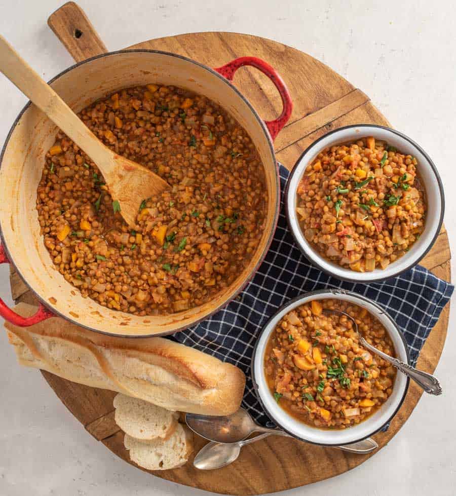 Round wooden platter with a pot and two bowls of vegetarian lentil soup and French bread on the side.