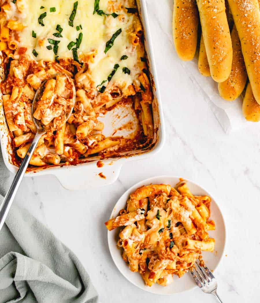 top view of a plate and baking dish of baked ziti with breadsticks on the side