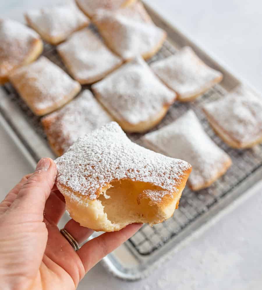 picture of a hand holding a beignet that has a bite out of it with beignets in the background