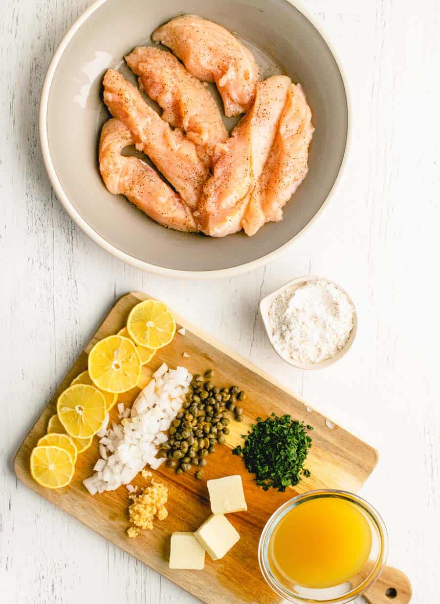 top view of a bowl of chicken and a cutting board with the ingredients for chicken picatta on it