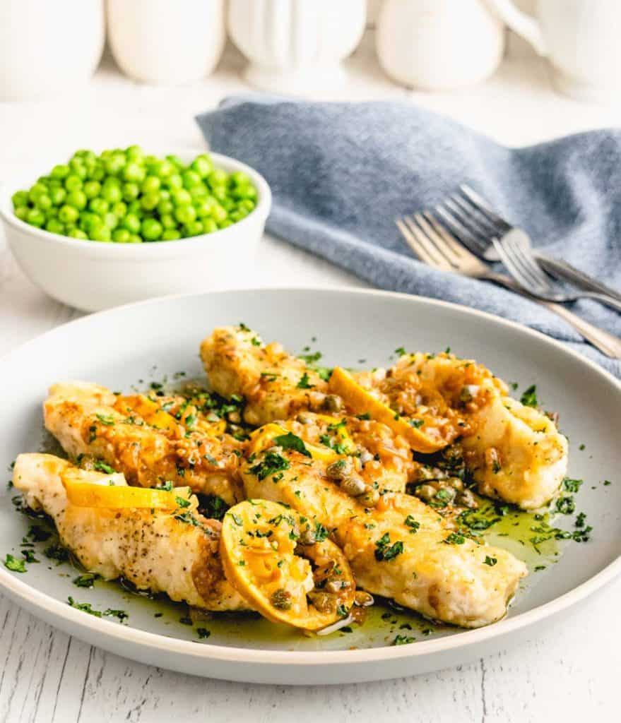 a plate of chicken picatta with a bowl of peas and forks in the background
