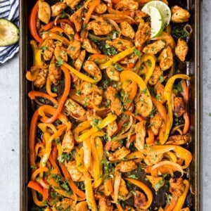 A baking sheet filled with cooked sheet pan chicken fajitas—juicy strips, red and yellow bell peppers, onions, all garnished with chopped herbs and lime wedges.