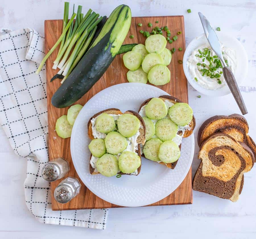 top view of a plate of cucumber sandwiches with the ingredients for them next to the plate