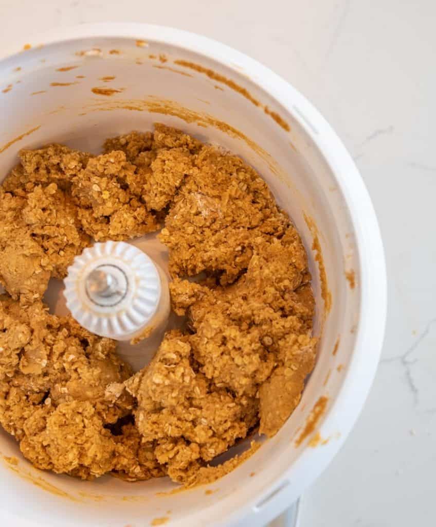 A food processor bowl containing a thick, light brown dough mixture pressed against the sides, viewed from above on a white countertop.
