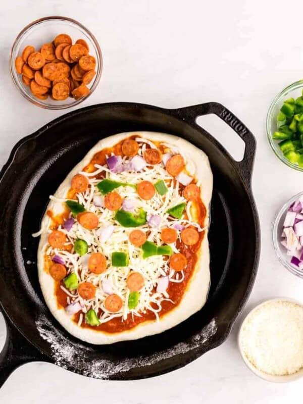 top view of uncooked pizza in a skillet with ingredients in bowls surrounding the skillet