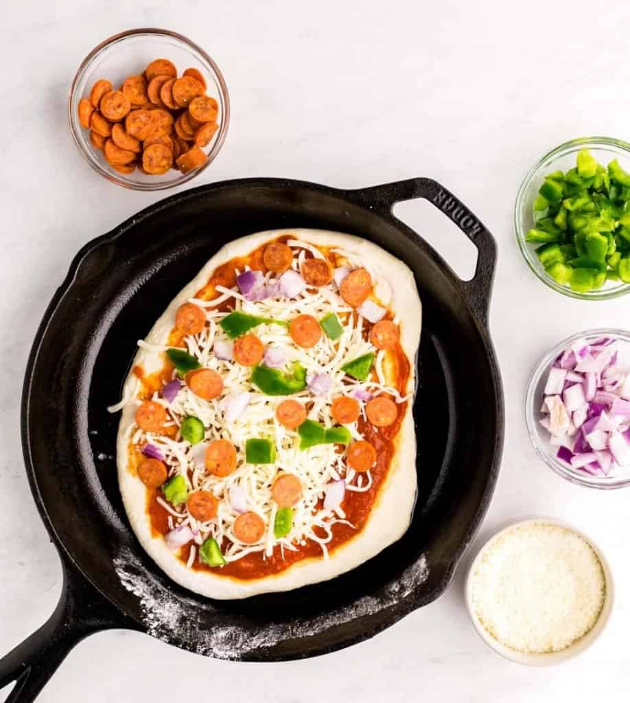 top view of uncooked pizza in a skillet with ingredients in bowls surrounding the skillet