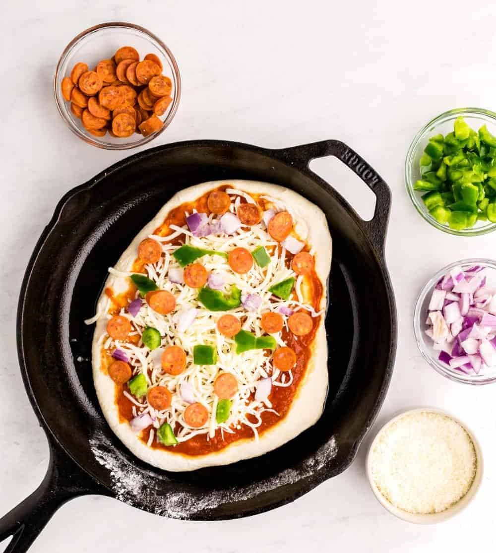 top view of uncooked pizza in a skillet with ingredients in bowls surrounding the skillet