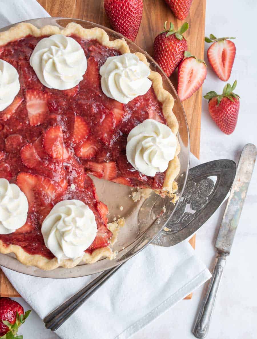 top view of a strawberry pie with dollops of whipped cream on it, with a slice missing.