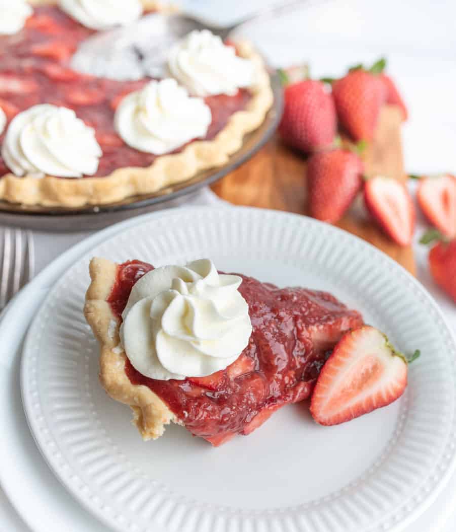 close up picture of a slice of strawberry pie on a plate with a strawberry pie in the background.