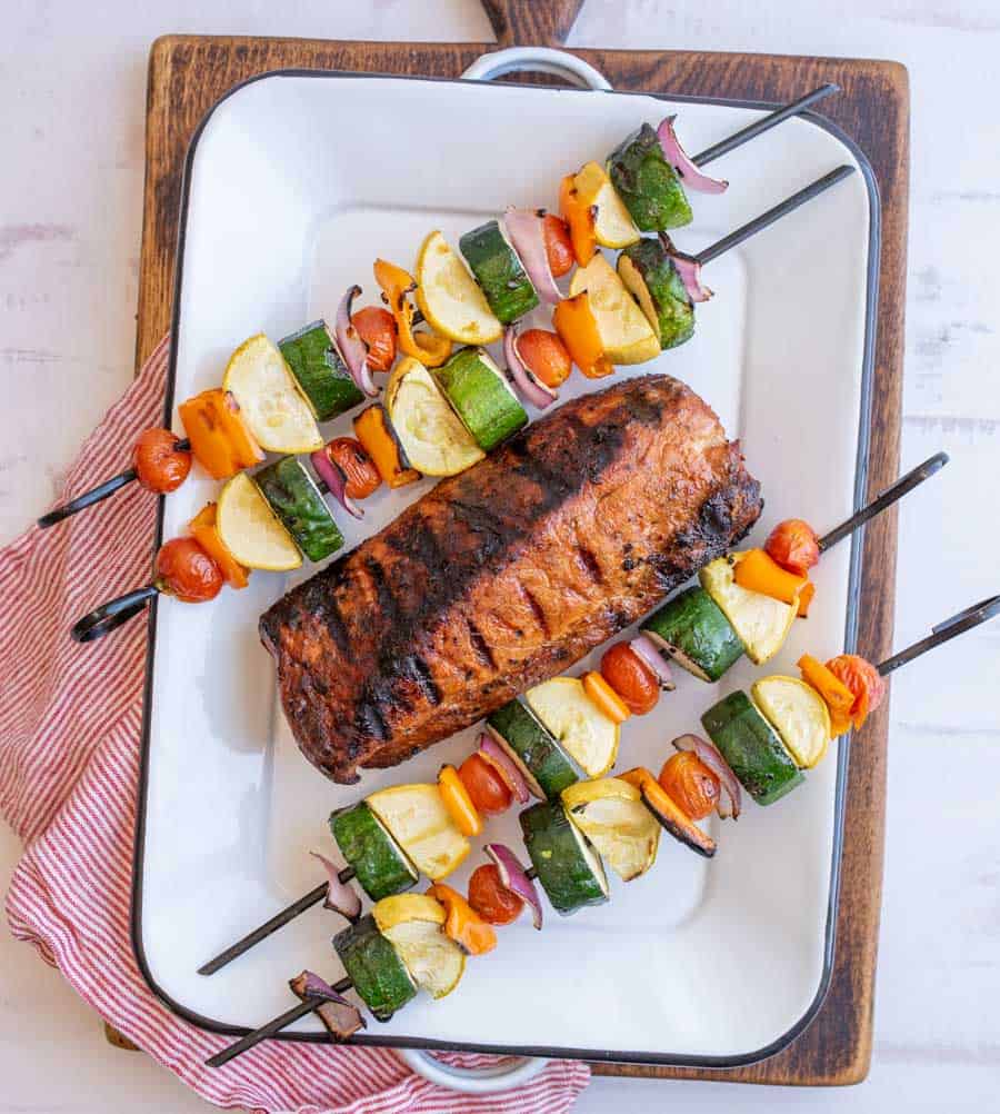 A white platter with a whole grilled pork loin filet at an angle in the middle with two grilled veggie kabobs on either side of it