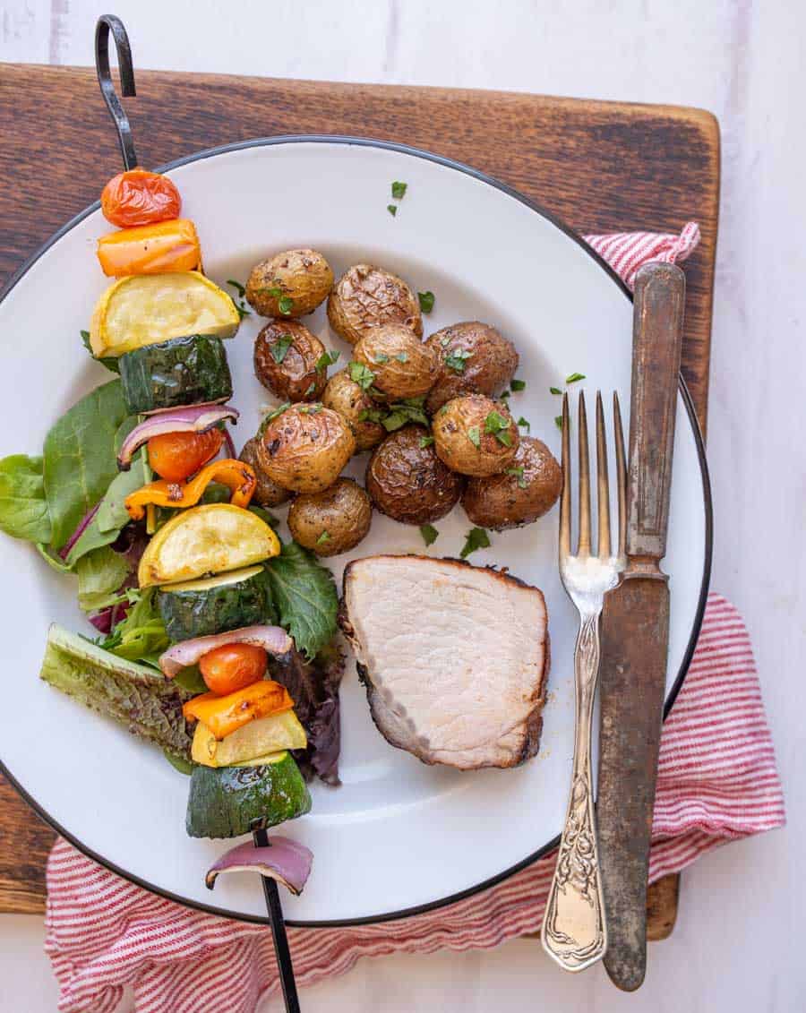 a white plate with a grilled veggie kabob, slice of grilled pork loin, and roasted potatoes