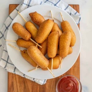 A white plate of mini corn dogs on sticks, made with a classic corn dog recipe, sits on a wooden board with a checkered towel underneath. A small bowl of ketchup is placed next to the plate.