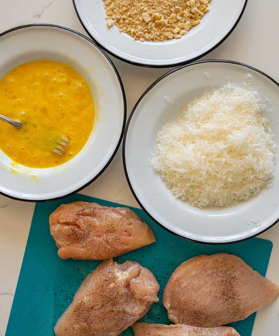 top view of ingredients for parmesan crusted chicken in bowls next to chicken breasts on a cutting board