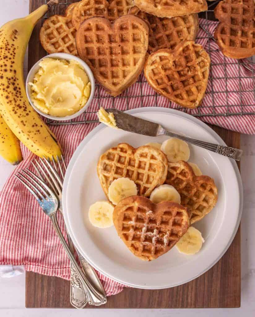 top view of heart shaped waffles and slices of bananas on a plate with utensils, bananas, bowl of butter, and other heart shaped waffles on a cooling rack