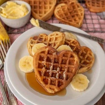 heart shaped waffles on a plate with syrup and banana slices and waffles in the background