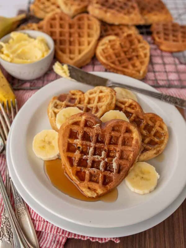 A plate with heart-shaped banana oatmeal waffles topped with sliced bananas and syrup. A bowl of butter sits beside the plate, with more waffles in the background on a checkered cloth. Silverware rests next to the plate on a wooden table.