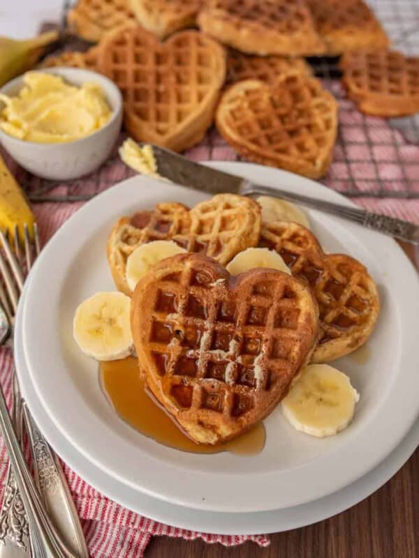 heart shaped waffles on a plate with syrup and banana slices and waffles in the background