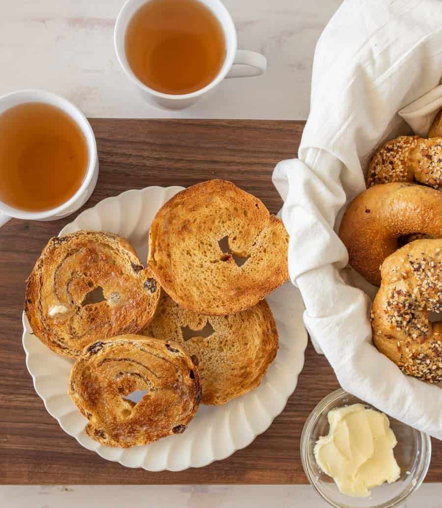 top view of bagels sliced and toasted on a plate next to two cups of tea and a bowl of bagels