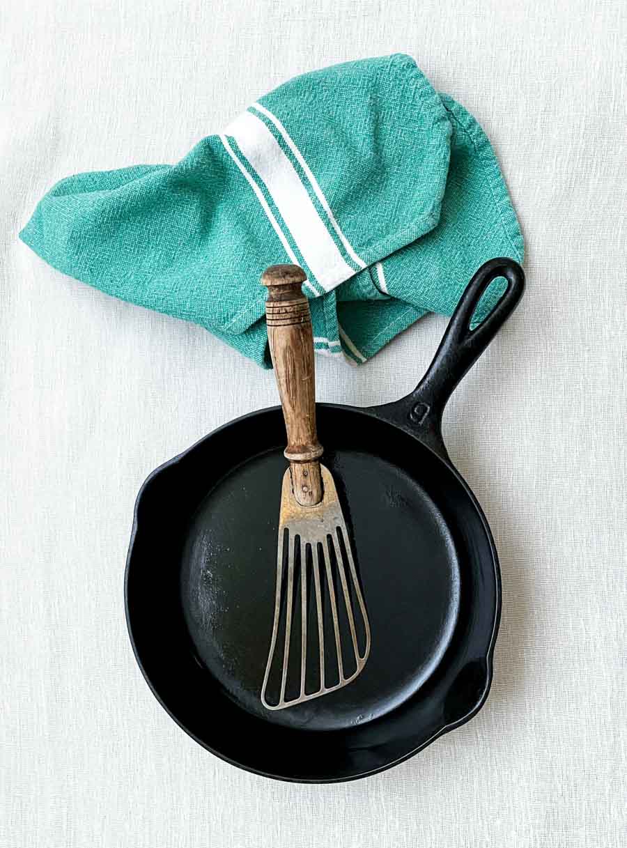 black cast iron skillet on a white background with a blue-green towel beside it and a metal spatula with brown wood handle in it.