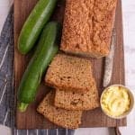 top view of a loaf of zucchini bread with some slices cut out of it next to zucchinis and a bowl of butter