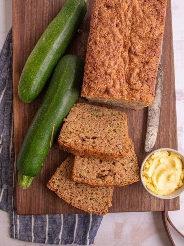 top view of a loaf of zucchini bread with some slices cut out of it next to zucchinis and a bowl of butter