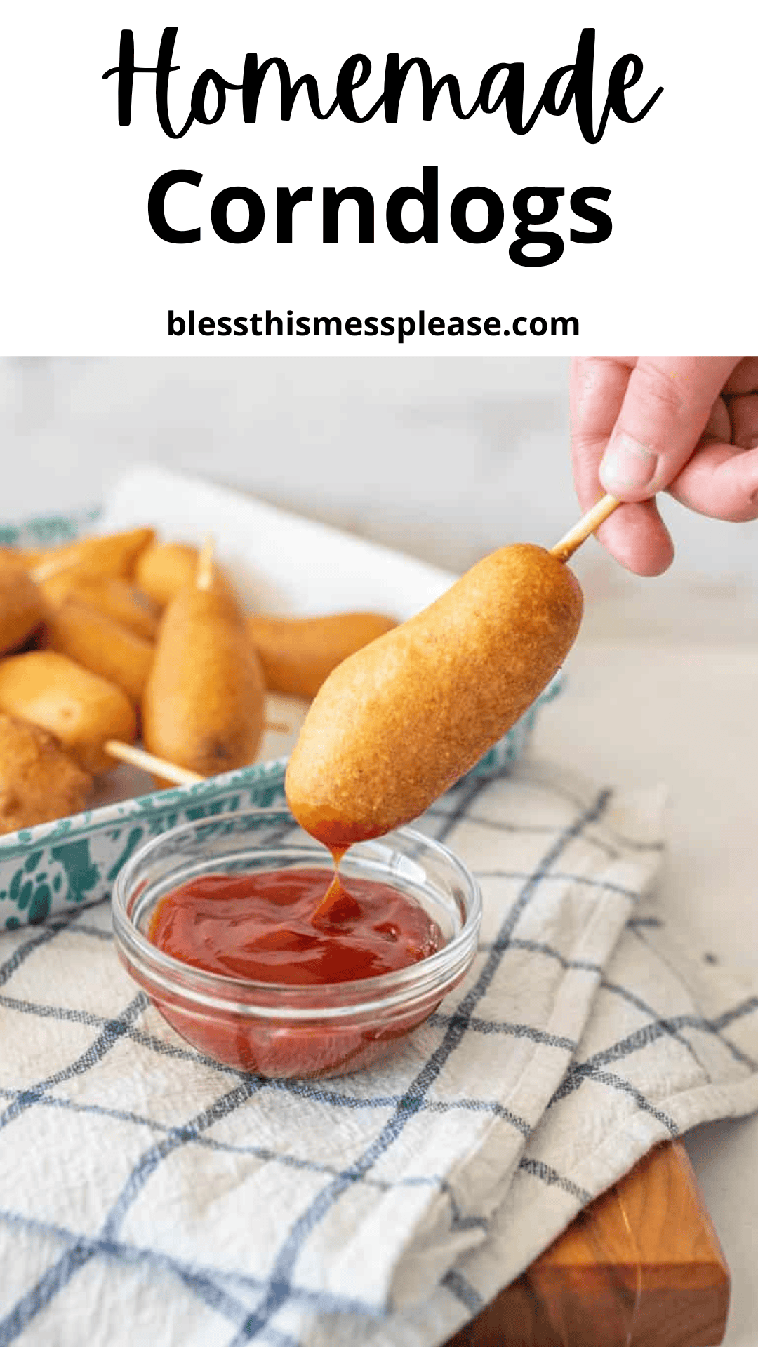 A hand dips a homemade corn dog into a small glass bowl of ketchup, with more corn dogs on a tray in the background. The text above reads Homemade Corndogs corn dog recipe blessthismessplease.com.