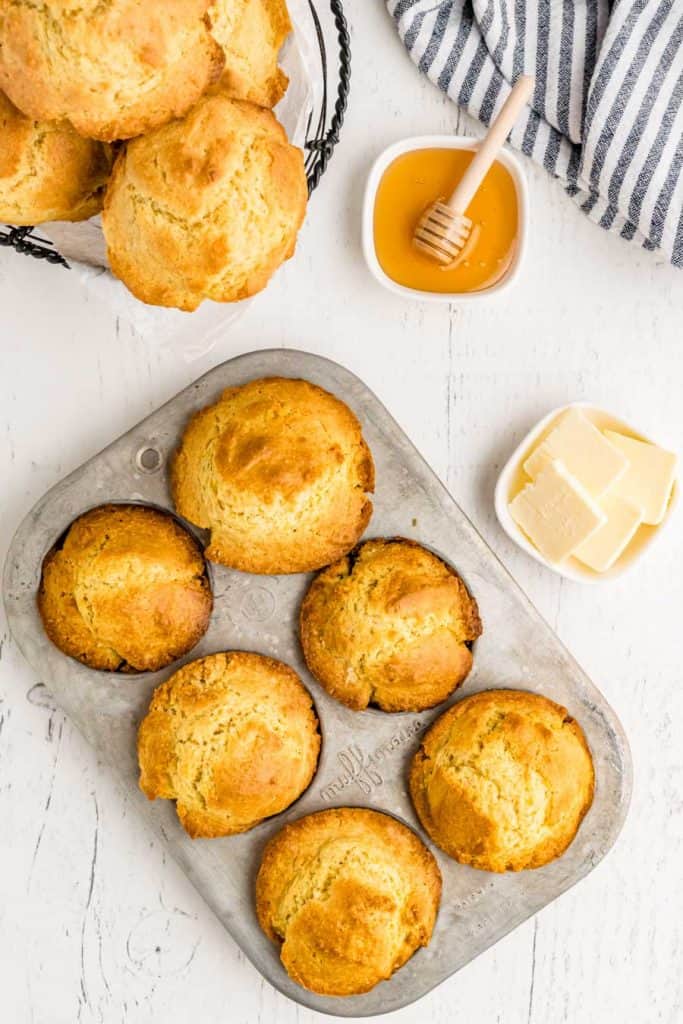 A muffin tin with six golden brown biscuits from a classic corn muffin recipe sits on a white table. Nearby are a wire basket with more biscuits, a bowl of honey with a dipper, a striped towel, and a dish of sliced butter.