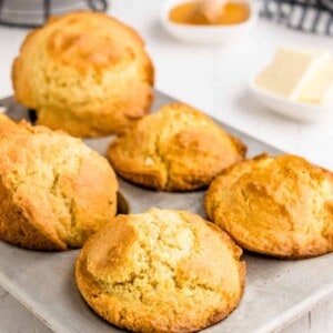 A muffin pan with five golden-brown muffins rests on a white surface, crafted from a classic corn muffin recipe. In the background are a small dish of butter, a bowl of honey with a dipper, and a striped towel.