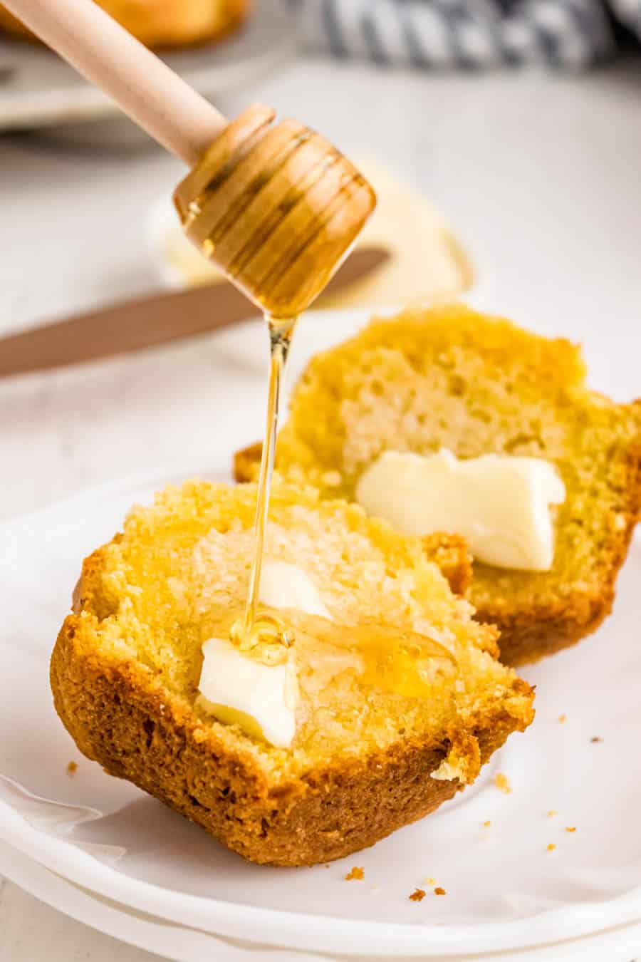 Halved corn muffin slathered with butter and topped with honey dripping from a honey stick.