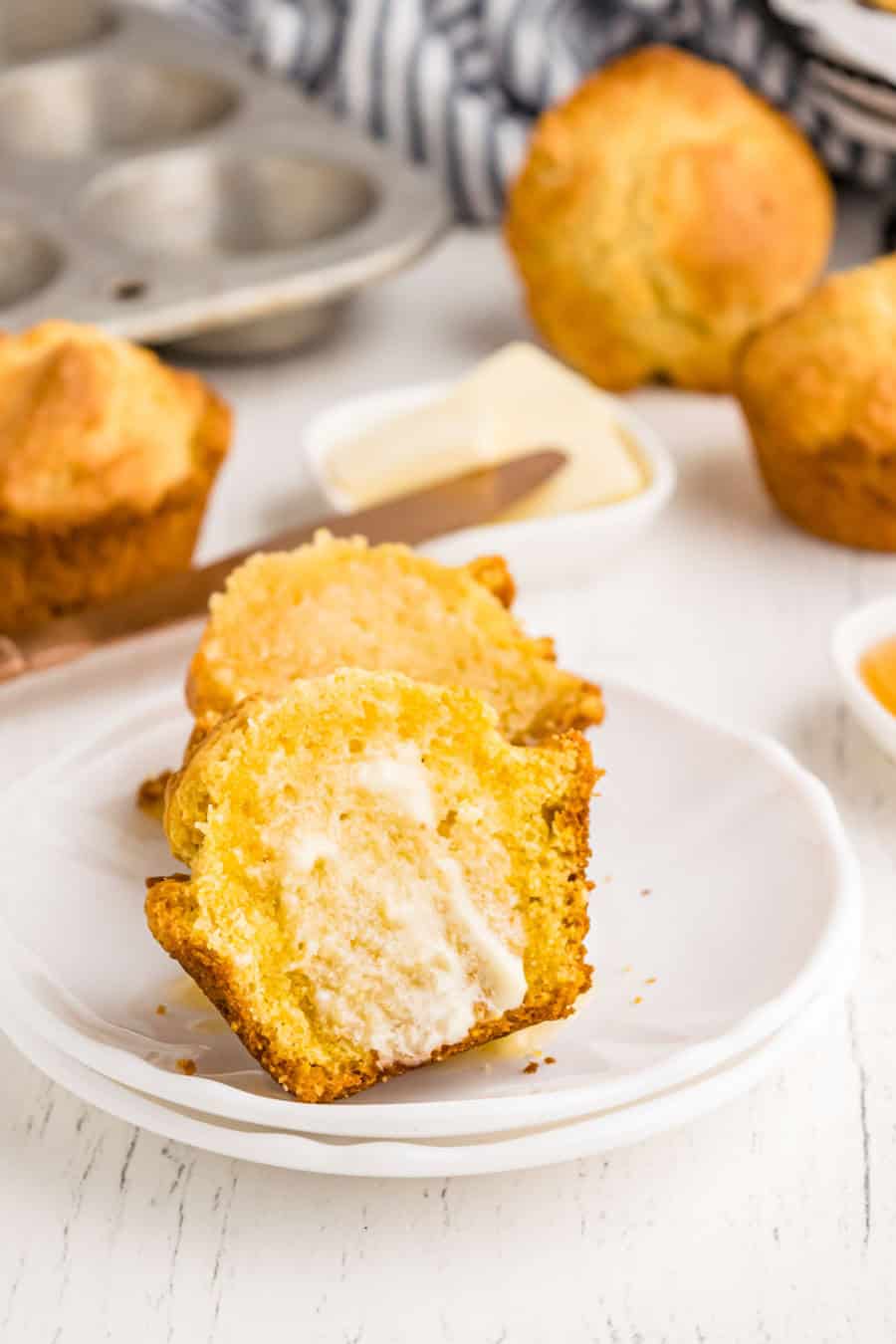 A golden cornbread muffin, made from a classic corn muffin recipe, is sliced open on a white plate with melted butter inside. More muffins, a muffin tin, and a butter dish appear in the background.
