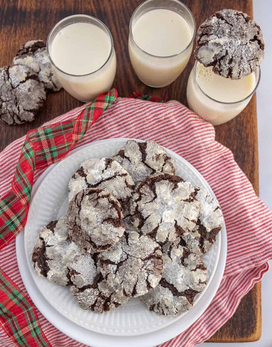 Top view of a white plate on a red striped napkin with chocolate crinkle cookies on top and glasses of milk next to it.