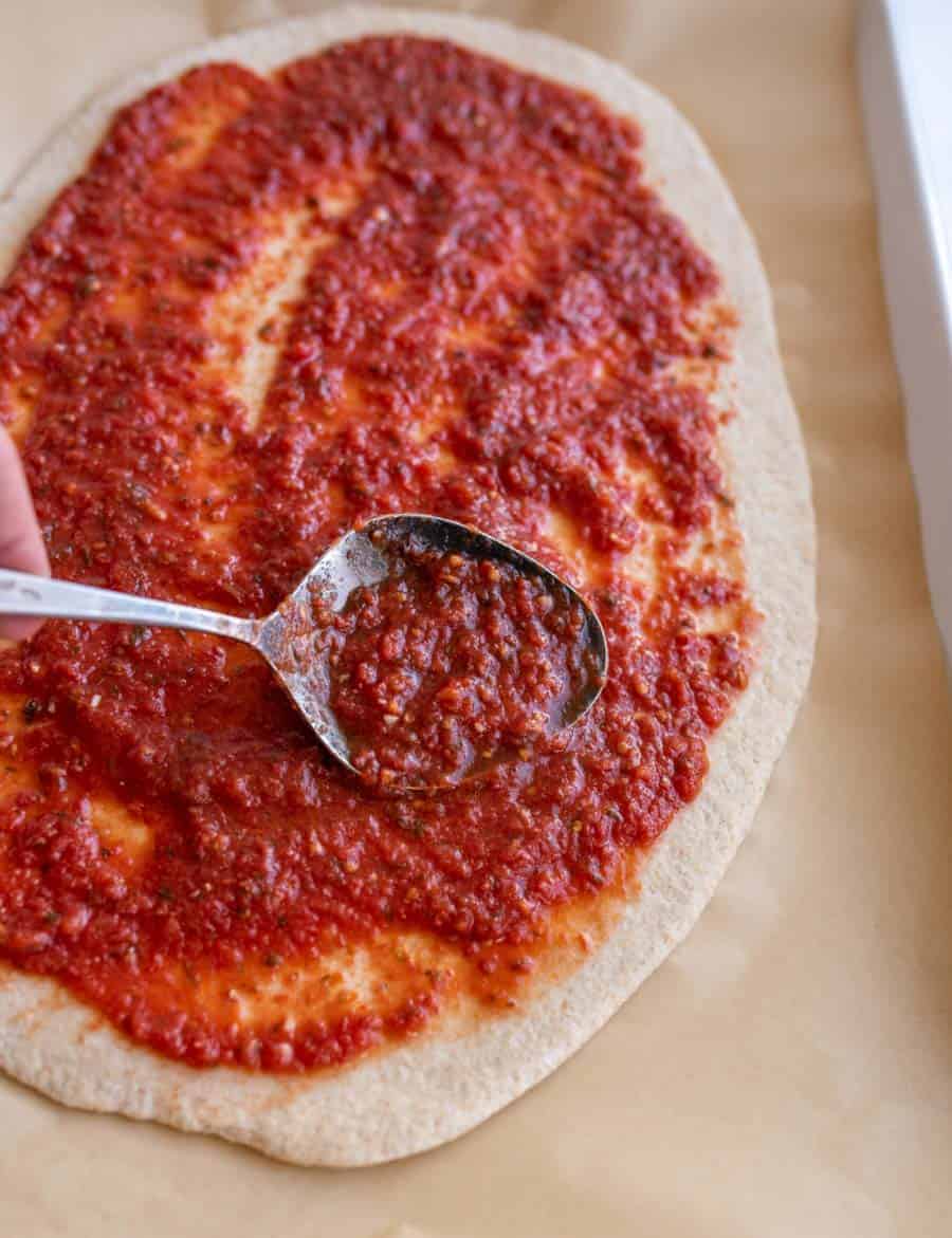 Pizza sauce being spread over pizza dough with a metal spoon.