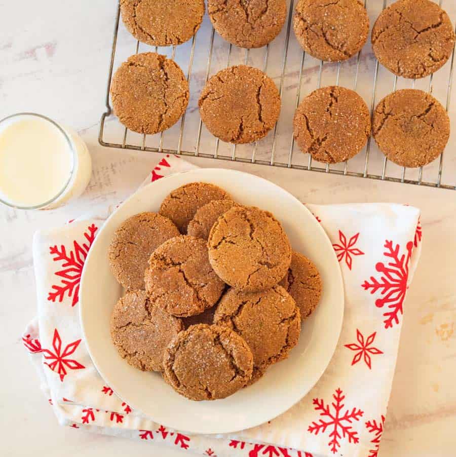 An arrangement of gingersnap cookies on a white plate atop a decorative white napkin with red snowflakes. A glass of milk and gingersnaps on a baking rack off to the side.