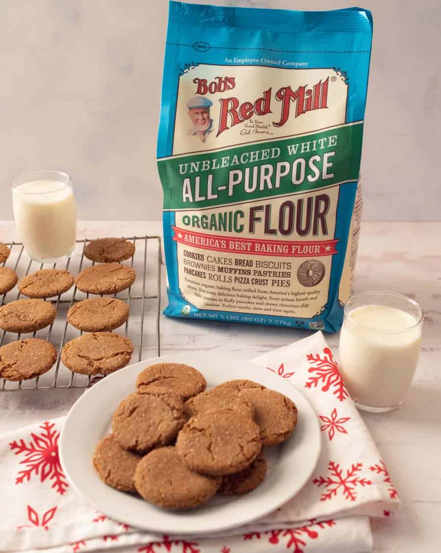 ginger snap cookies on a white plate and baking rack with glasses of milk and a bag of flour