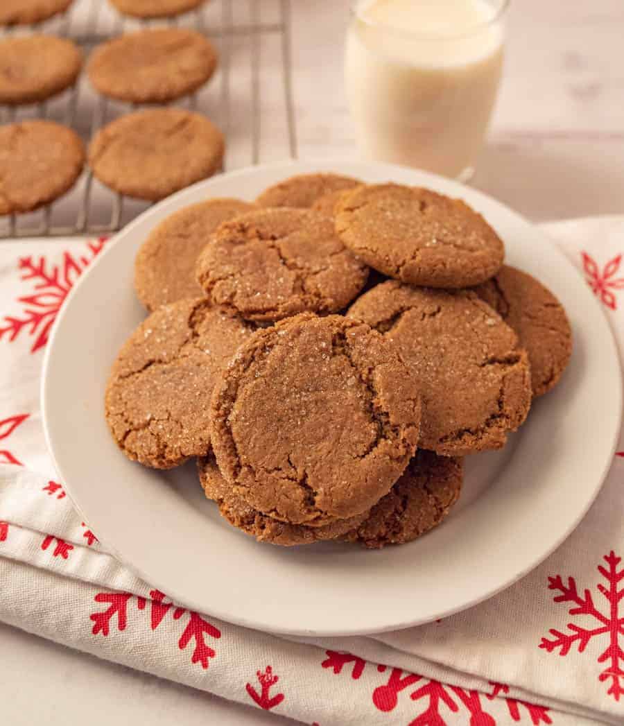 A plate of gingersnap cookies on a white plate on a white linen with red snowflakes and a glass of milk.