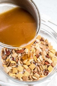 carmel sauce being poured from a pan into a clear glass bowl of chex-mix.