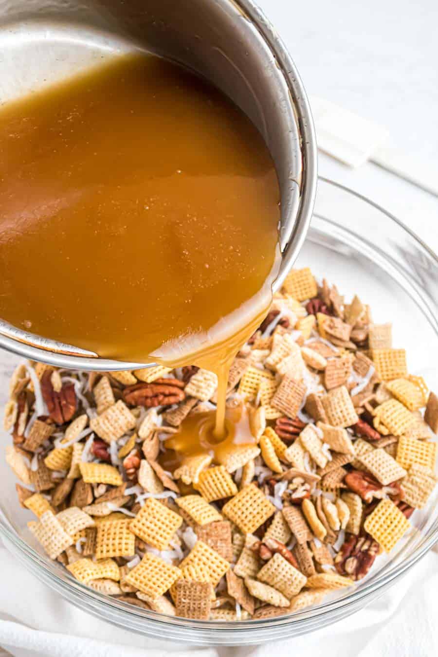 carmel sauce being poured from a pan into a clear glass bowl of chex-mix.